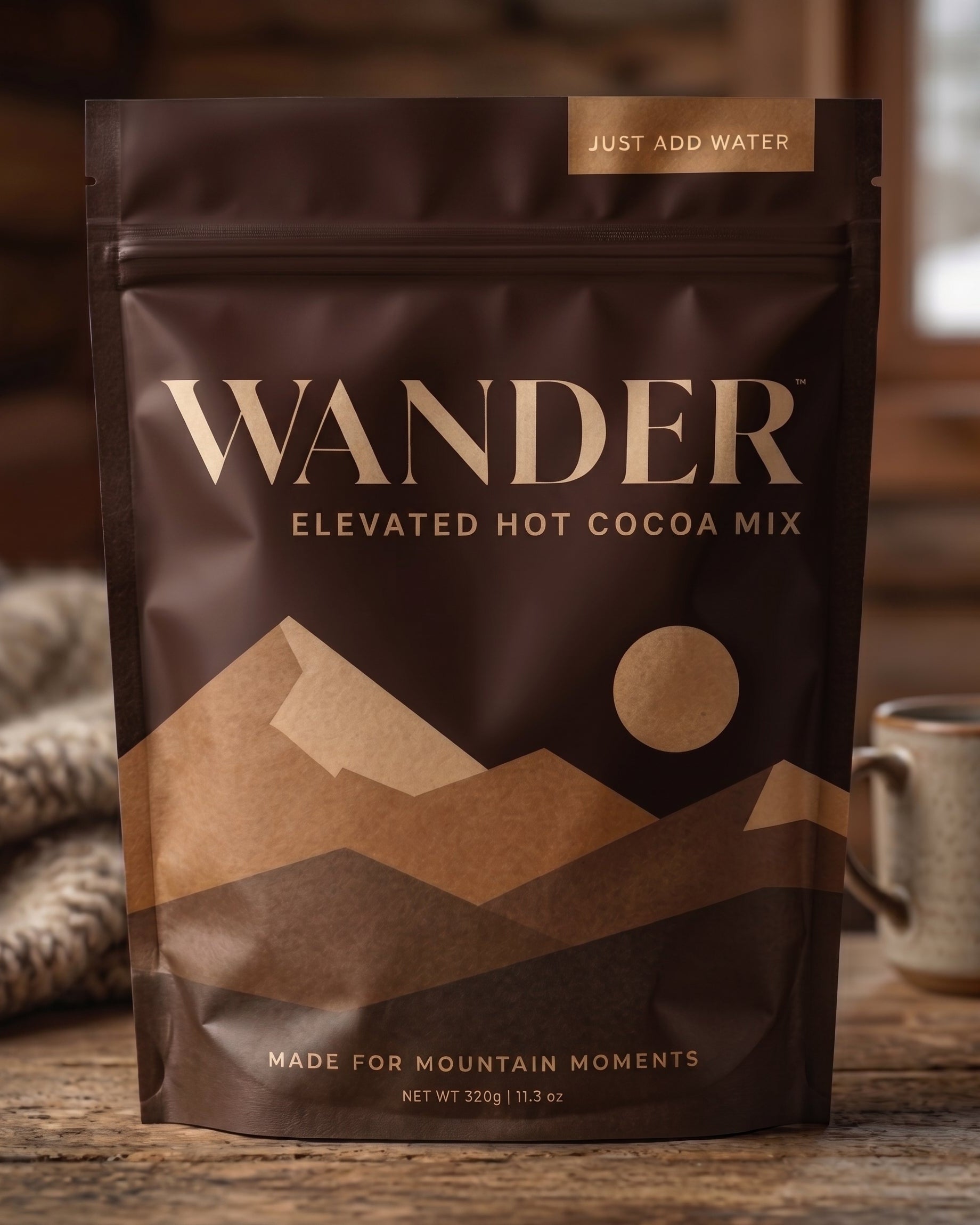 A stand-up pouch of Wander Elevated Hot Cocoa Mix on a rustic wooden surface, with a mug and soft indoor background visible around it.