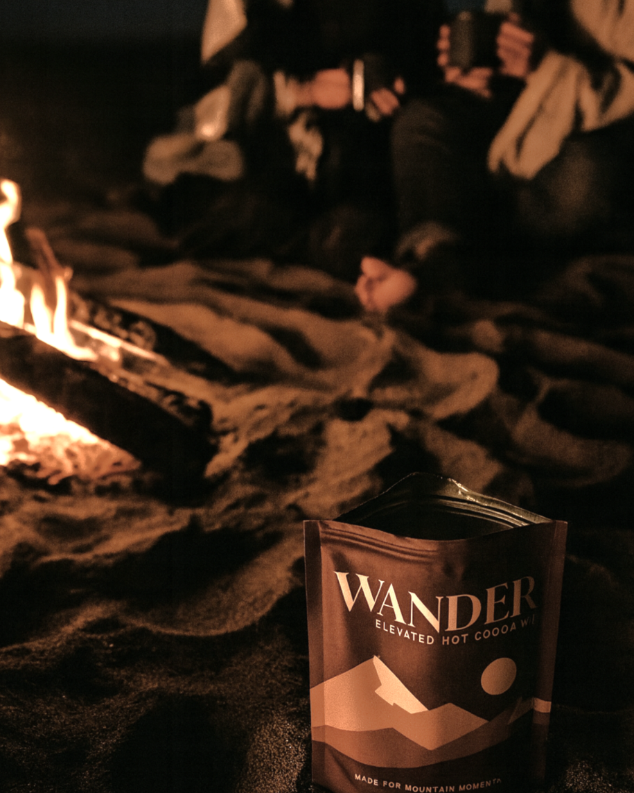 A WANDER Hot Cocoa Bean bag sits on dark sand in the foreground, with a campfire burning in the background and two people sitting and holding mugs in a warm, low-light outdoor setting.