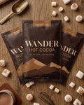 Three bags of Wander Hot Cocoa in milk chocolate flavor arranged on a wooden tabletop, surrounded by marshmallows, a cup of cocoa, a spoon, and a whisk. The product packaging is dark brown with gold text and mountain graphics.