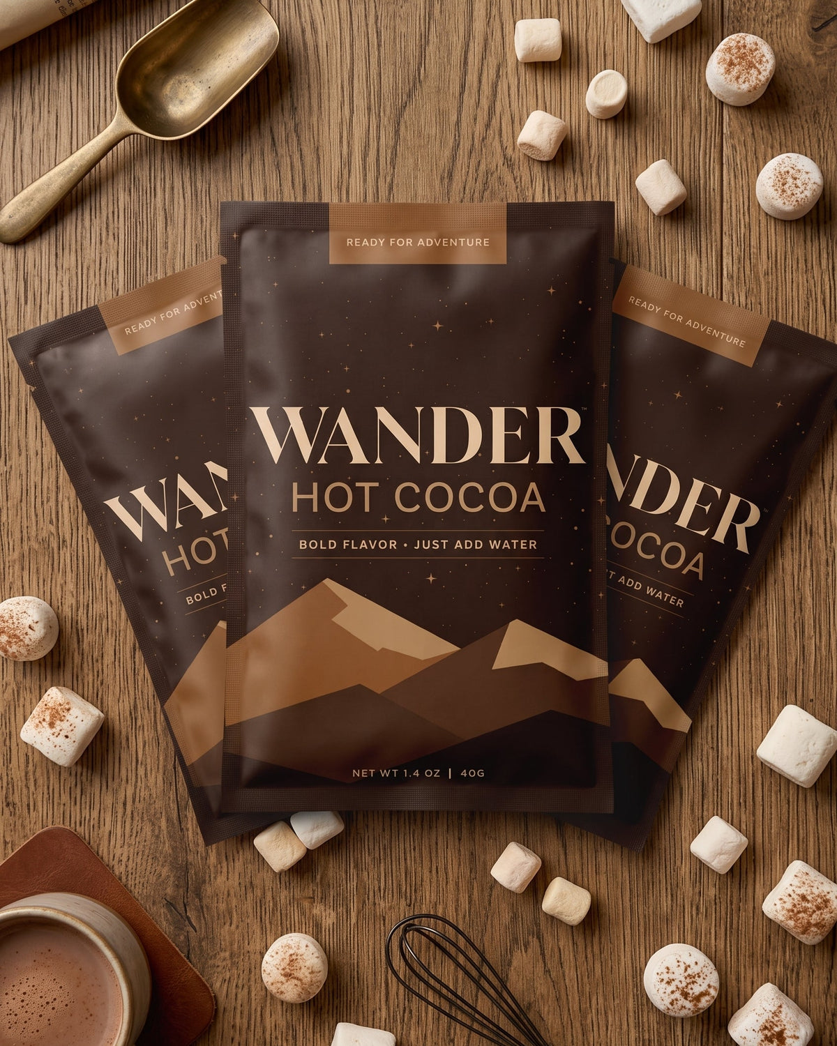 Three bags of Wander Hot Cocoa in milk chocolate flavor arranged on a wooden tabletop, surrounded by marshmallows, a cup of cocoa, a spoon, and a whisk. The product packaging is dark brown with gold text and mountain graphics.