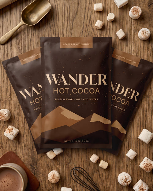 Three bags of Wander Hot Cocoa in milk chocolate flavor arranged on a wooden tabletop, surrounded by marshmallows, a cup of cocoa, a spoon, and a whisk. The product packaging is dark brown with gold text and mountain graphics.